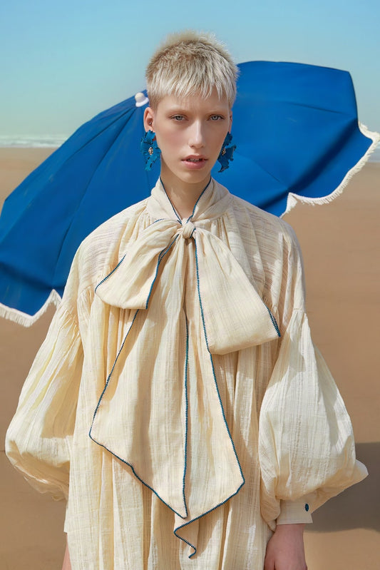 Woman wearing a beige dress with a large bow in a desert setting with a blue parasol.