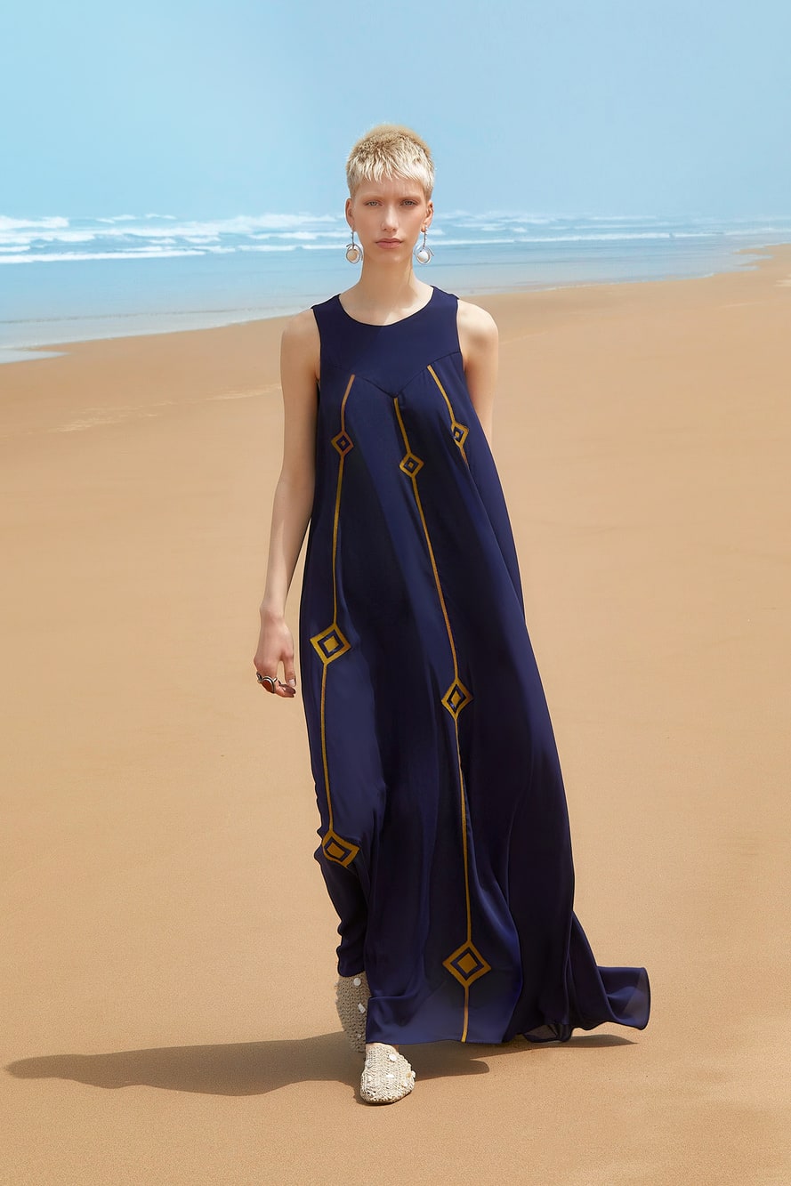 Woman in a long blue dress with geometric patterns standing on a sandy beach.